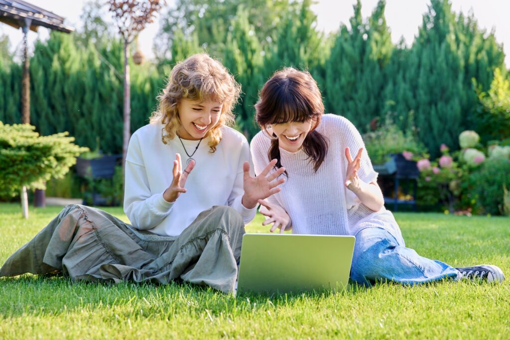Teenage female student friends laughing sitting on grass with laptop